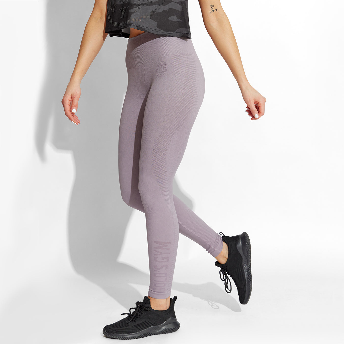 photo of female model wearing train seamless legging in purple. small golds gym weight plate logo near left hip. golds gym printed near left ankle vertically.