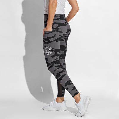 rear photo of female model wearing train high waist legging in black camo. large golds gym weight plate logo in white on left leg in middle below pocket. model posing with hands in side pockets.