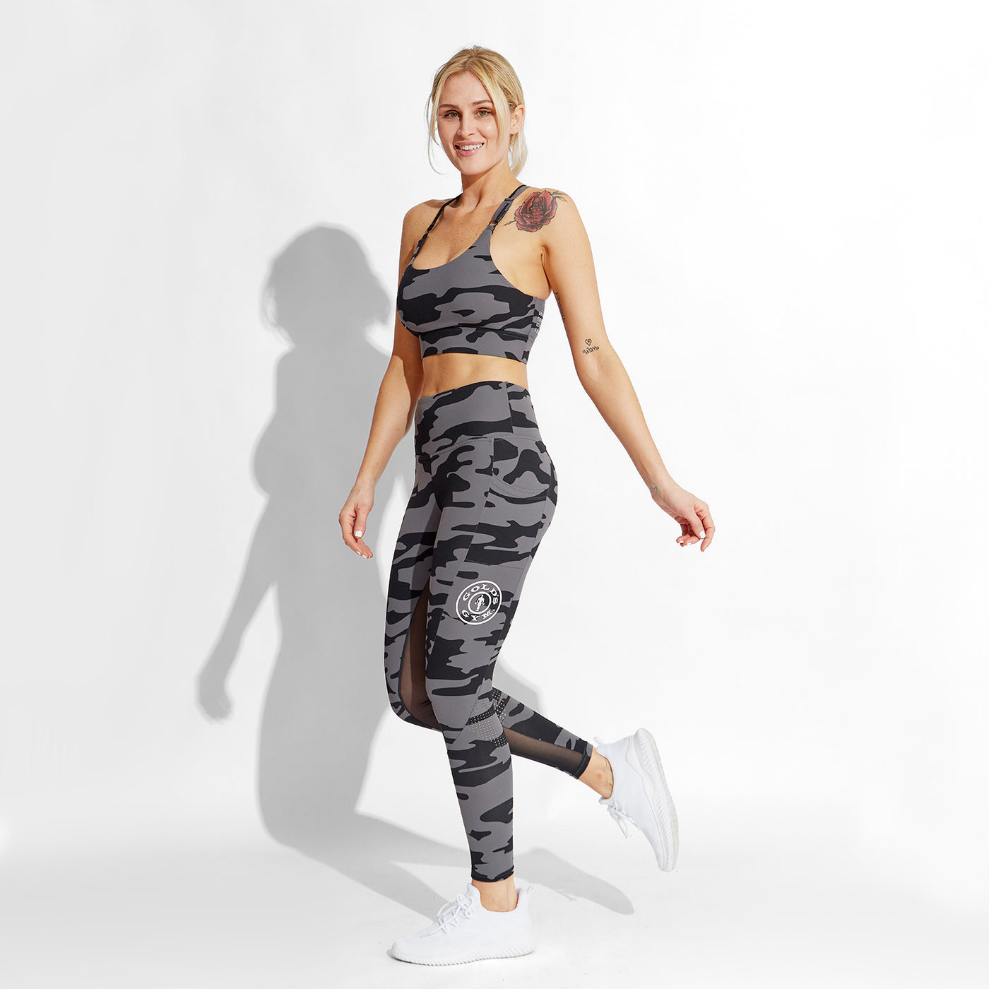 photo of female model wearing train high waist legging in black camo. large golds gym weight plate logo in white on left leg in middle below pocket.