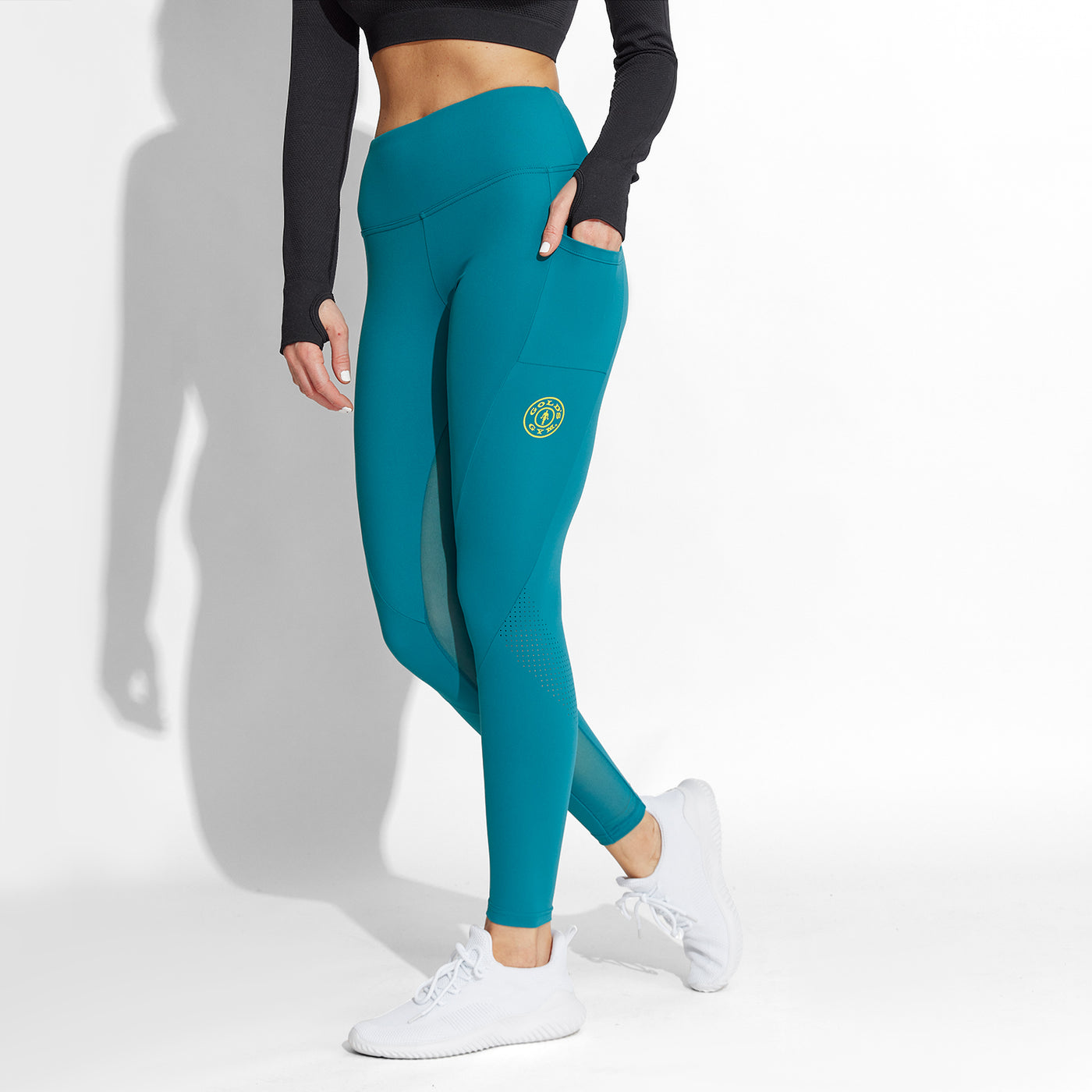 photo of female model wearing train waist legging in blue. small golds gym weight plate logo in yellow on left leg in middle below pocket. model posing with left hand in pocket